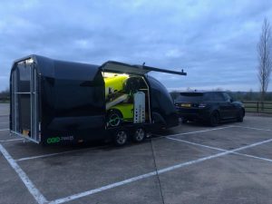 Black enclosed vehicle trailer with two neon yellow caterhams inside attached to a black range rover in a carpark