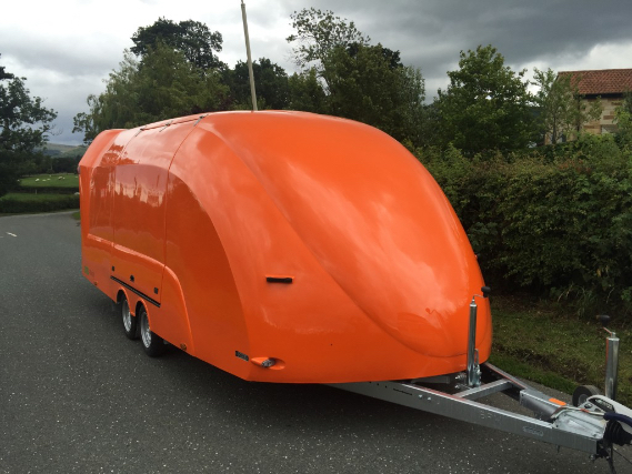 Orange enclosed vehicle trailer on a road in front of a hedge